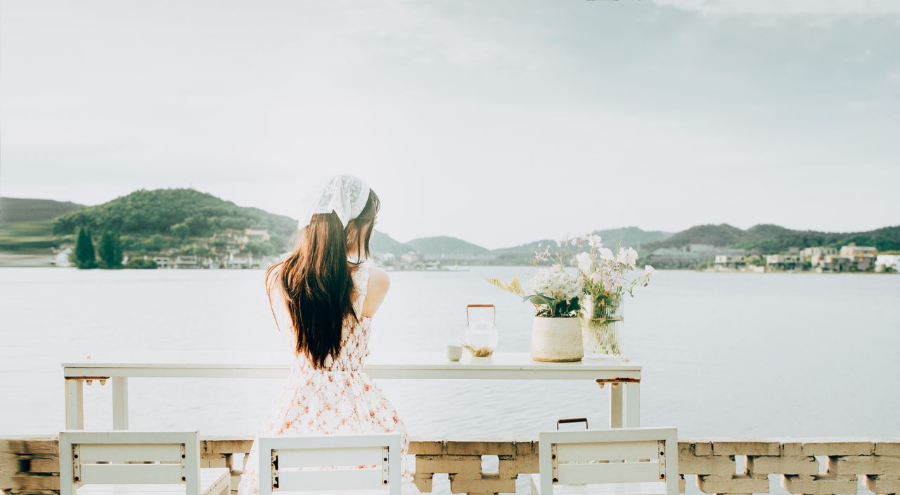 Home A woman with long hair and a floral dress sits by a table with vases by a serene lake.