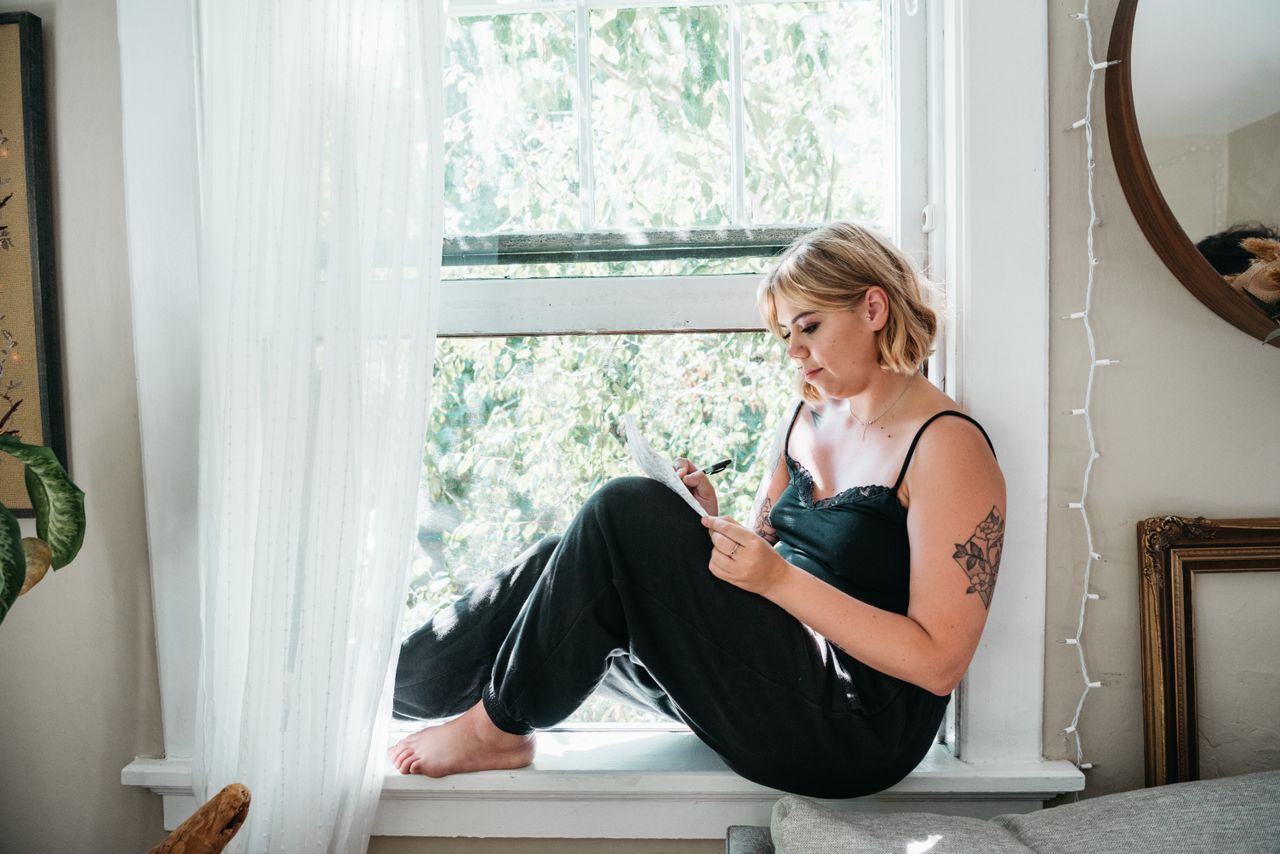 Home A young woman sits in a windowsill surrounded by natural light, writing in a notebook, creating a serene indoor scene.